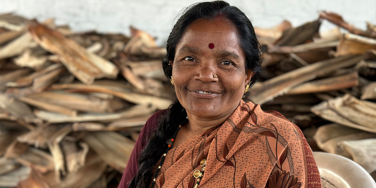 A middle-aged woman smiles at the camera. In the background is a pile of dried areca leaves.