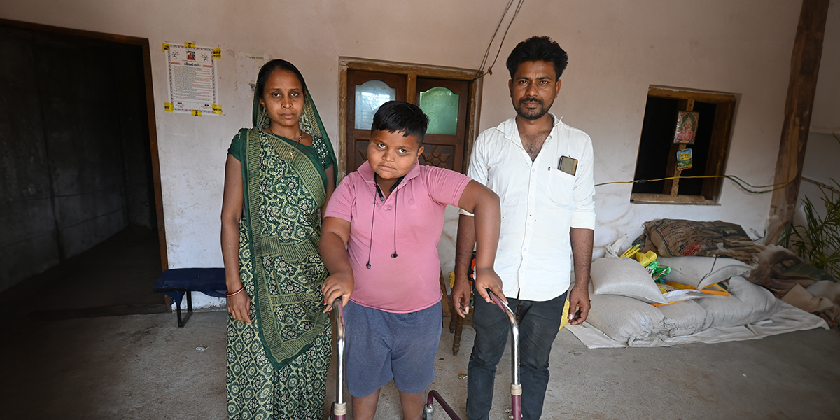 A boy standing with the help of a rollator between his parents. Behind then is their house.