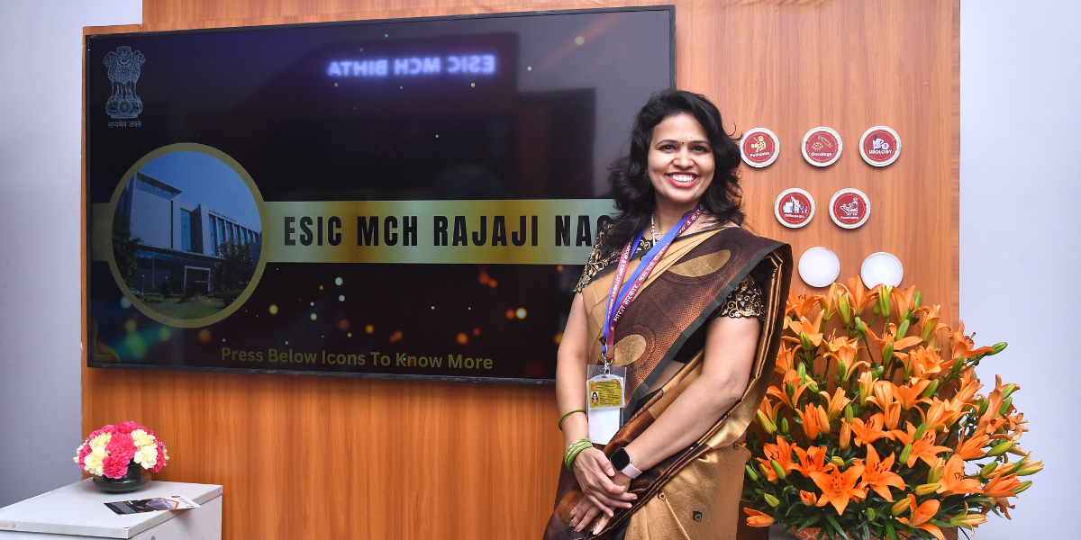 A smiling woman wearing a saree stands in front of a backdrop with a board reading 'ESIC MCH RAJAJI NAGAR'