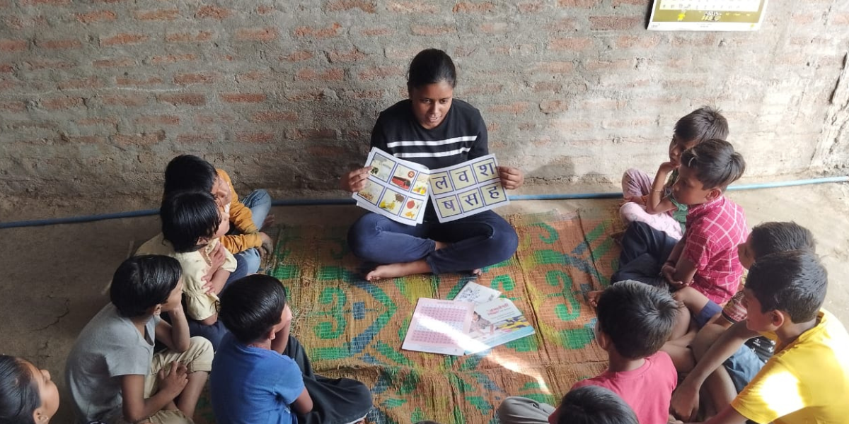 A young woman in her 20s, sits on a floor mat and teaches a group of children.