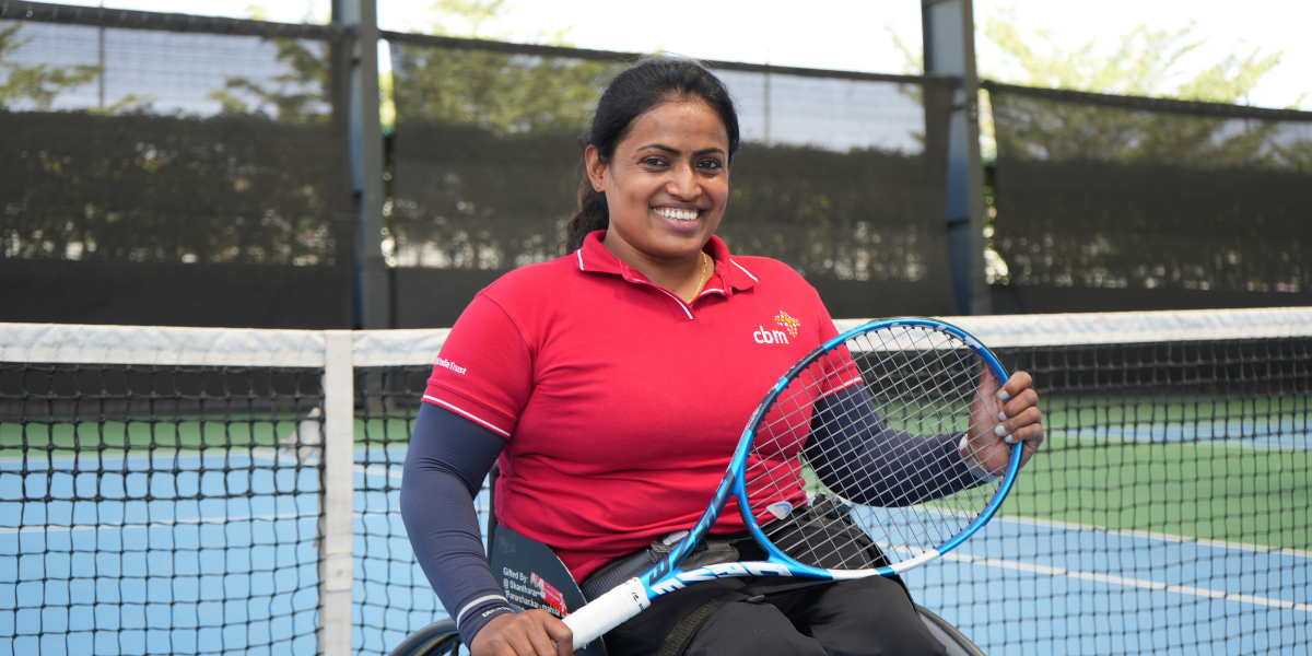 Shilpa, a woman in her 30s, smiles at the camera. Shilpa is sitting in a wheelchair and is holding a tennis racket. The photo is taken at a tennis court.