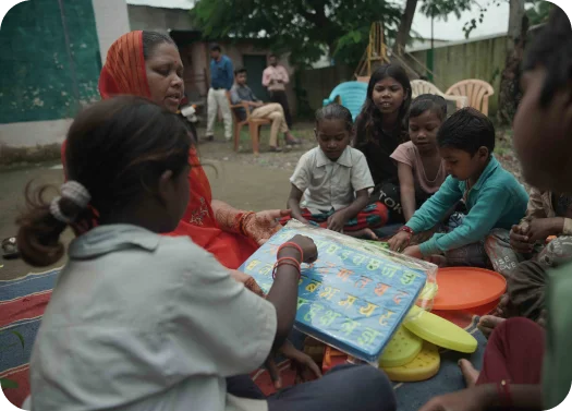 Children playing board game outdoors with adults seated in the background.