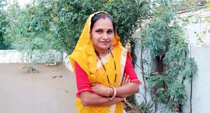 Person in a yellow and red traditional outfit with folded arms, standing beside greenery.