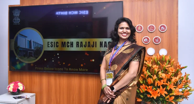 A person in traditional attire stands in front of a sign for ESIC MCH RAJAJI NAGAR with flowers on either side.