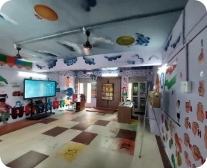 A colorful classroom with educational wall decorations, patterned floor, and a hanging TV.