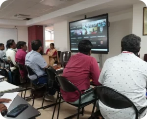 A group of people in a meeting room watching a presentation on a large screen.