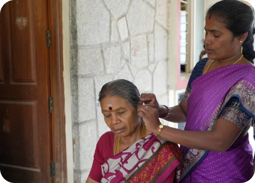 Two women in traditional Indian attire, one standing and adjusting the other's earring.