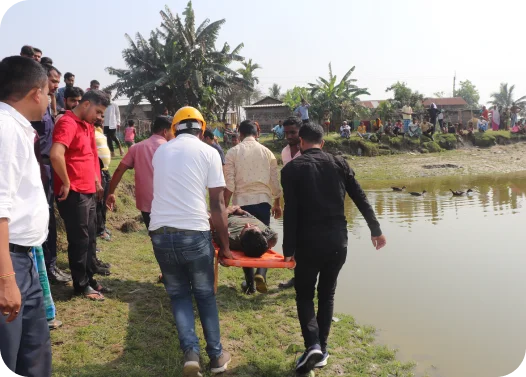 People carrying someone on a stretcher near a pond, with onlookers standing by.