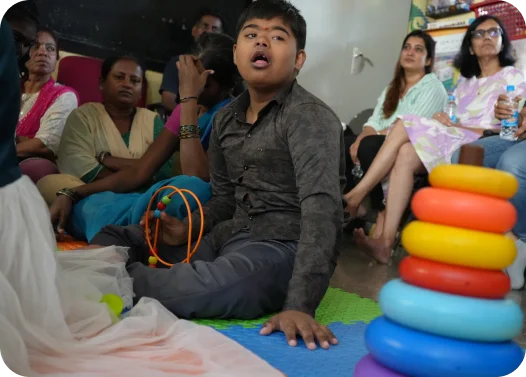 A group of people sitting inside a room with colorful play mats and stacked toy rings on the right.