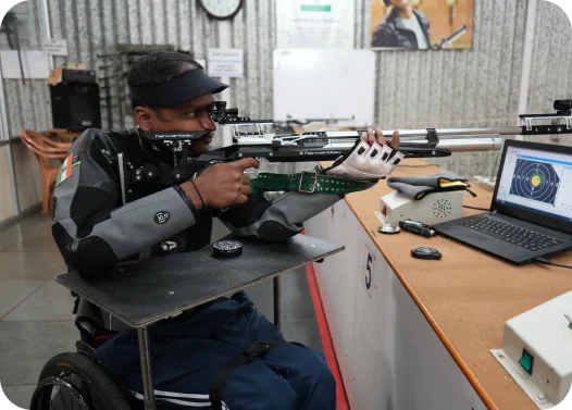 Person in a wheelchair at a shooting range aiming a rifle, with a target displayed on a laptop screen.