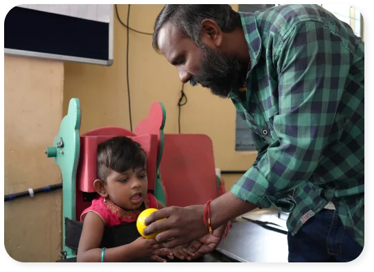 An adult holding a child's hands over a table, with a colorful chair in the background.