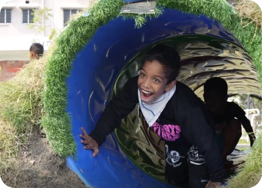 Individuals on a playground, one emerging from a blue tunnel slide lined with grass.